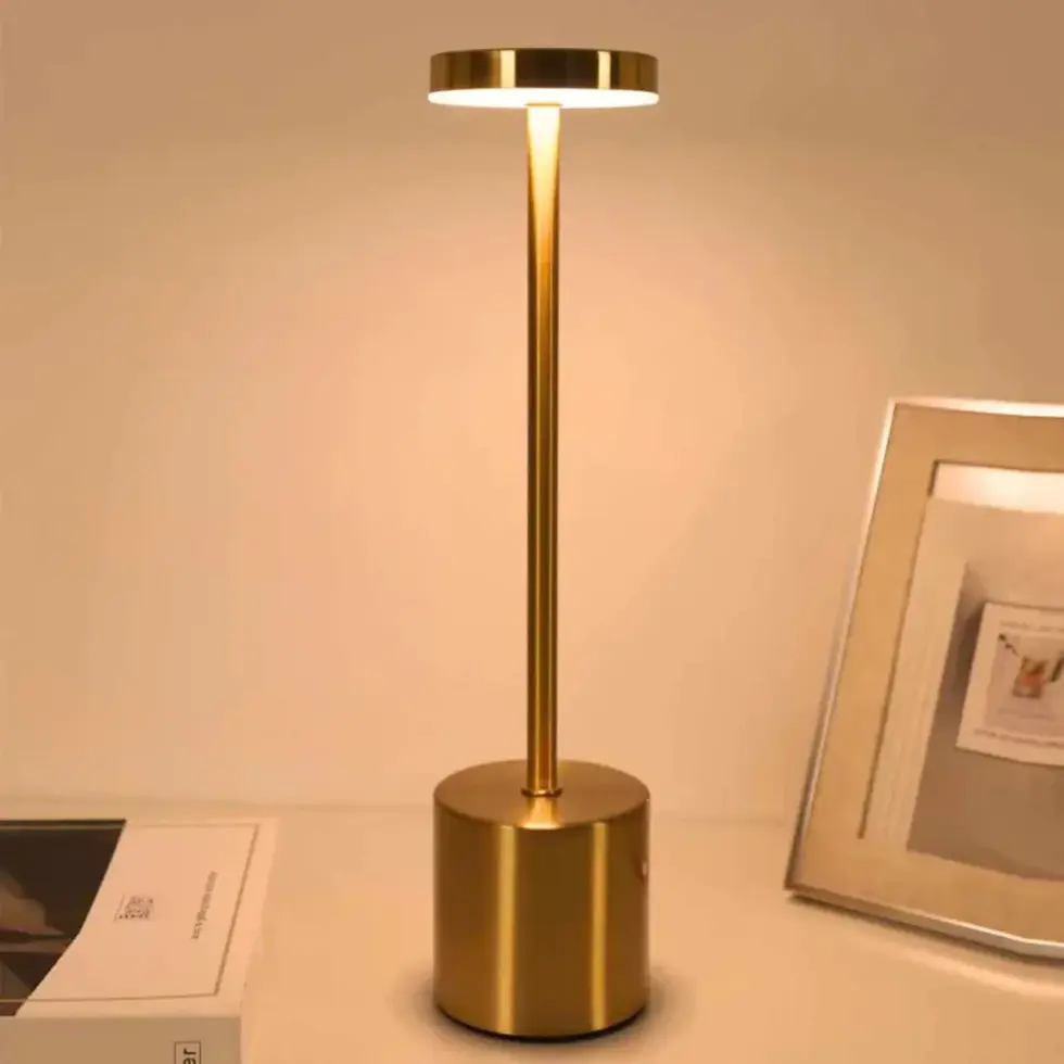 Luminária dourada acesa sobre mesa bege, ao lado de livro e porta-retratos. Ambiente minimalista e aconchegante, tons quentes.