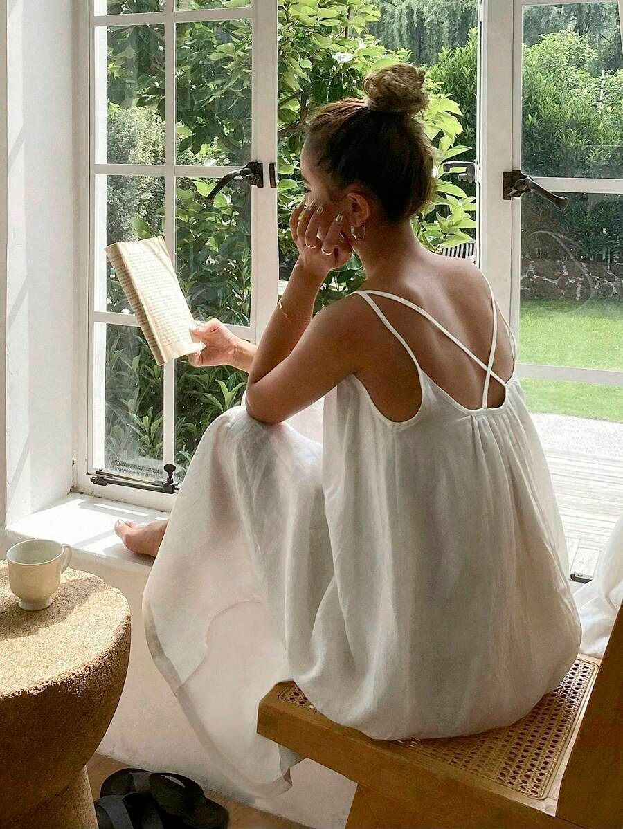 Mulher de vestido branco lê junto à janela, sentada, com jardim verde ao fundo e xícara ao lado, clima calmo.