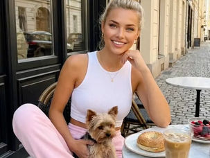 Mulher sorridente com cão no colo em café ao ar livre, com café gelado, pão e frutas, numa rua de pedra.