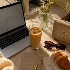 Mesa ensolarada com laptop, café gelado, flores, diário, croissant, óculos e bolsa YSL; notas com texto manuscrito.