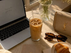 Mesa ensolarada com laptop, café gelado, flores, diário, croissant, óculos e bolsa YSL; notas com texto manuscrito.