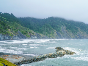 Rocky Lost Coast Trail with waves crashing, green cliffs shrouded in fog. A lone car on the shore. Moody, serene seascape.