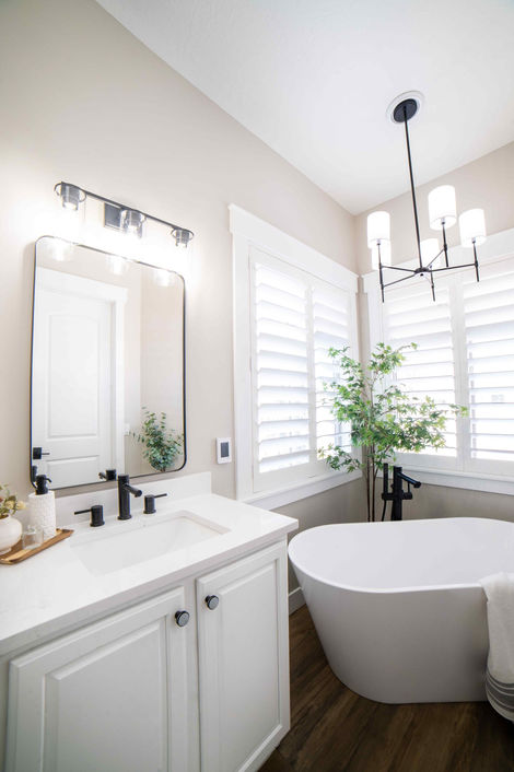 a bright attractive lavish bathroom with white vanity, free standing white bathroom tub, a chandelier & a plant in the corner