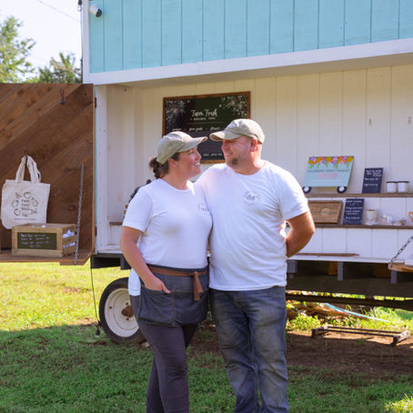 A man and a woman standing in front of an outdoor market stand with their arms around one another smiling at each other
