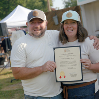 A man and a woman hold up a citation award in front of an outdoor market