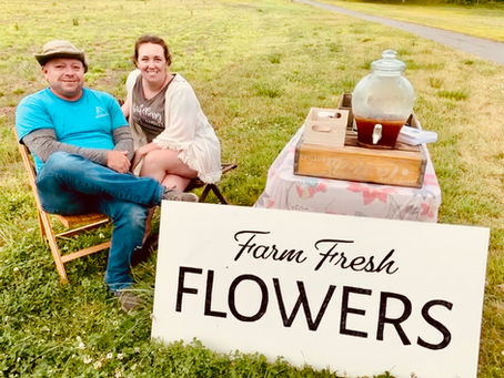 A man and a woman sitting on chairs in a grassy field at a small table with a sign that says "Farm Fresh Flowers"