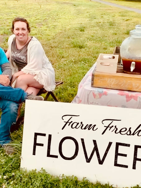 A man and a woman sitting on chairs in a grassy field at a small table with a sign that says "Farm Fresh Flowers"
