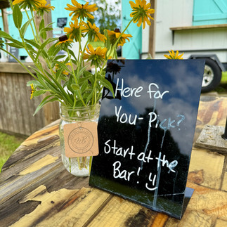 Flowers in a jar on a wooden table next to a sign that says "Here for you pick? Start at the bar!"