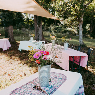 Tables with multicolored table cloths in an outdoor setting with buckets of flowers on the tables