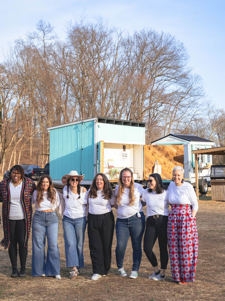 Group of people wearing white shirts and smiling, standing outdoors in a field with bare trees. A small blue and wood structure is in the background.
