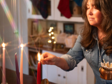 A woman lights a red candle with a match. in the background there are stockings, lights and other Christmas decorations.