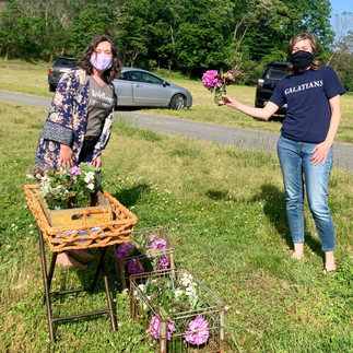 Two women standing far apart with masks on in a field. One woman holds flowers