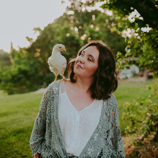 A woman stands in a field with a chicken on her shoulder