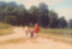 Three children walking hand in hand on a dirt path in a sunny, wooded area, with one in a red coat. The mood is nostalgic and serene.