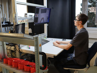 Eine Person sitzt an einem technischen Arbeitsplatz mit zwei Monitoren, die ein Assistenzsystem anzeigen. Auf dem Tisch stehen Werkzeuge und Behälter mit Kleinteilen, im Hintergrund ein heller Raum mit Regalen und Fenstern.