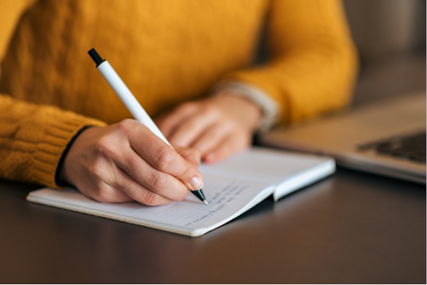 Eine Person in gelbem Pullover schreibt mit einem Stift in ein offenes Notizbuch, neben einem aufgeklappten Laptop.