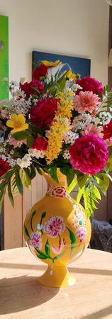Boeket met grote gele vaas met fushia pioenrozen, cosmea, lichtroze Gerbera's en eucalyptus. De vaas staat in een mooi daglicht. Het is een luxe kunstboeket die je kan huren