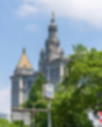 City Hall building with ornate spires against blue sky, green trees below. NYPD security camera sign in foreground, conveying a secure mood.