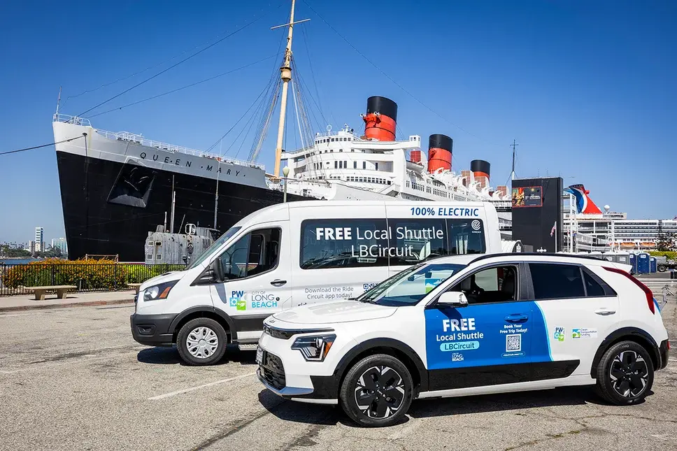 Two Circuit shuttle vehicles, a shuttle bus and a sedan, parked by the Queen Mary ocean liner i Long Beach, Calif. under a clear blue sky.