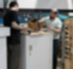 Two men handle a large drone on a workbench in an industrial setting. Background features Valqari logo and a wooden crate.