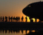 Silhouette of soldiers boarding a U.S. Air Force plane at sunset. Orange sky and reflection in water create a dramatic scene. Calm mood.