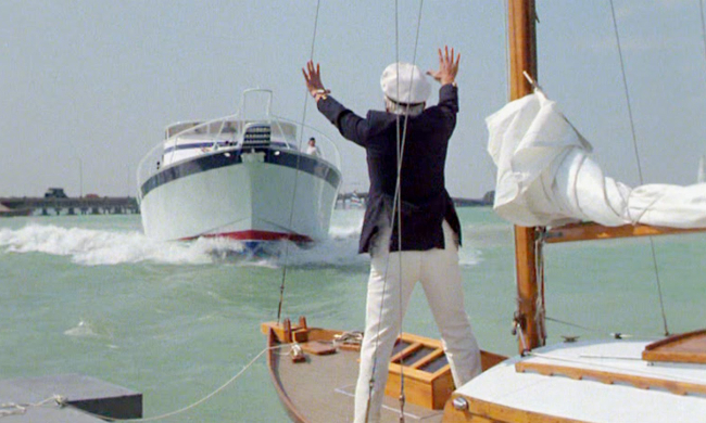 A person in a white cap and navy jacket raises both hands on a sailboat as a motorboat approaches.
