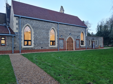 Stone building with arched windows and wooden doors, set against a grassy field and clear sky. Peaceful, evening lighting.