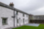 White stone cottage with green-trimmed windows, slate patio and lawn under a gray sky, quiet rural scene.