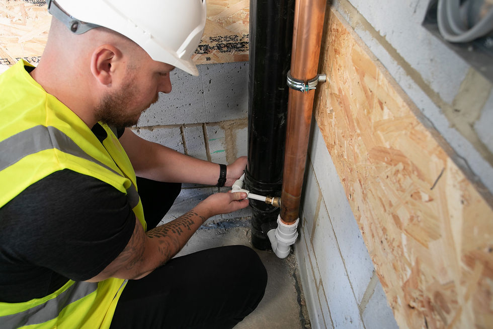 A construction worker in a white helmet and yellow vest fixes a pipe in a concrete and wood-paneled corner. Focused and engaged.