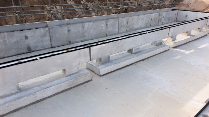 Concrete drainage channel with raised side walls and open slots along a sunlit construction site, no people visible