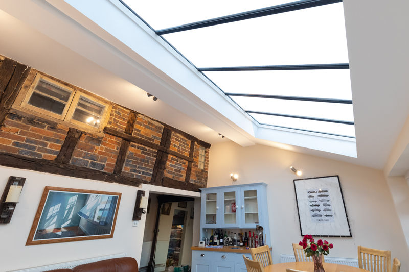 Room with glass ceiling, exposed brick, and wooden beams. Blue cabinet with bottles, dining table with flowers. Bright and airy ambiance.