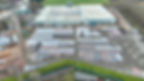 Aerial view of an industrial complex with a large warehouse, stacks of materials organized neatly outside. Overcast sky and green fields in the background.