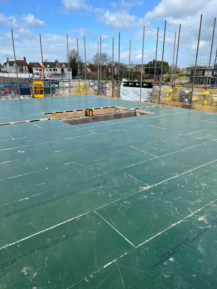 Construction site with green floor decking and steel poles, fenced off with houses and blue sky in the background.
