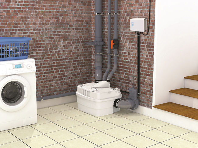 Laundry room with white washing machine and blue basket. Brick wall with pipes and a small box. Tiled floor and wooden stairs nearby.