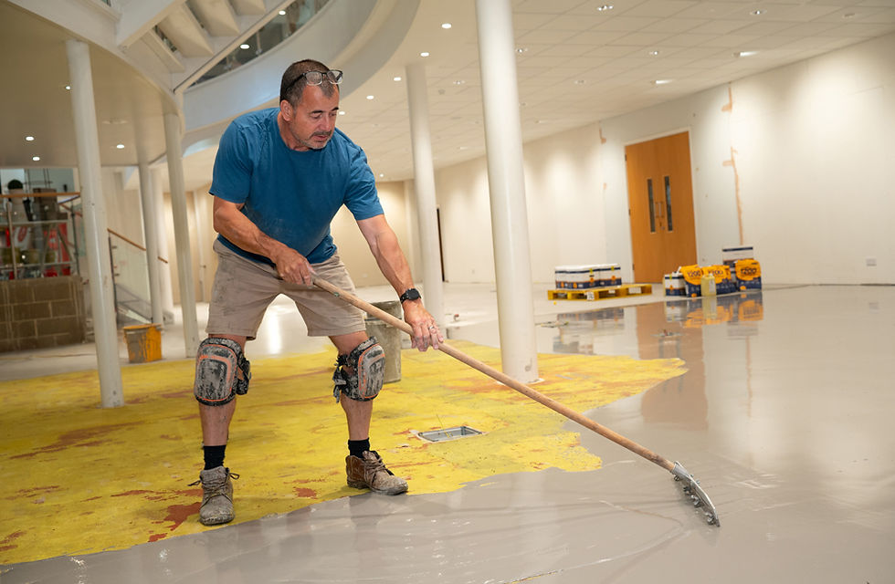 Man in blue shirt and knee pads smoothing grey floor in a building under renovation. Yellow patches and construction tools visible.