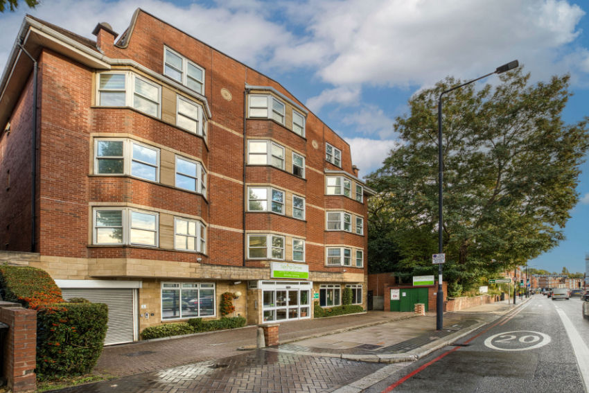 Red brick apartment building with green Spring Grove sign beside a tree-lined street and 20 mph road marking.