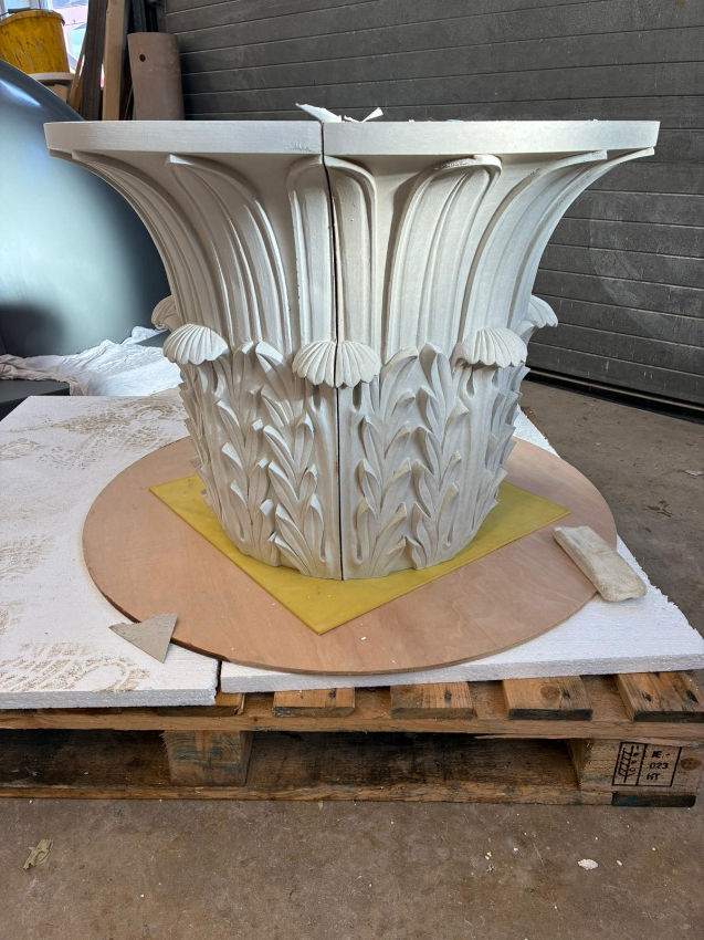 Unfinished white carved architectural column capital on a pallet in a workshop, with floral leaf relief and a visible center seam