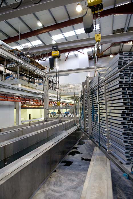 Industrial interior with metal beams on large racks, hoisted by yellow cranes. Overhead fluorescent lights illuminate the space.