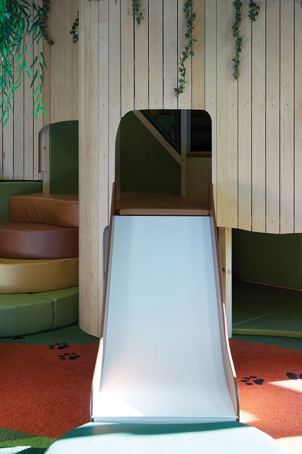 Children's play area with a wooden structure, white slide, and colorful padded steps. Greenery decor on walls; paw prints on the orange floor.