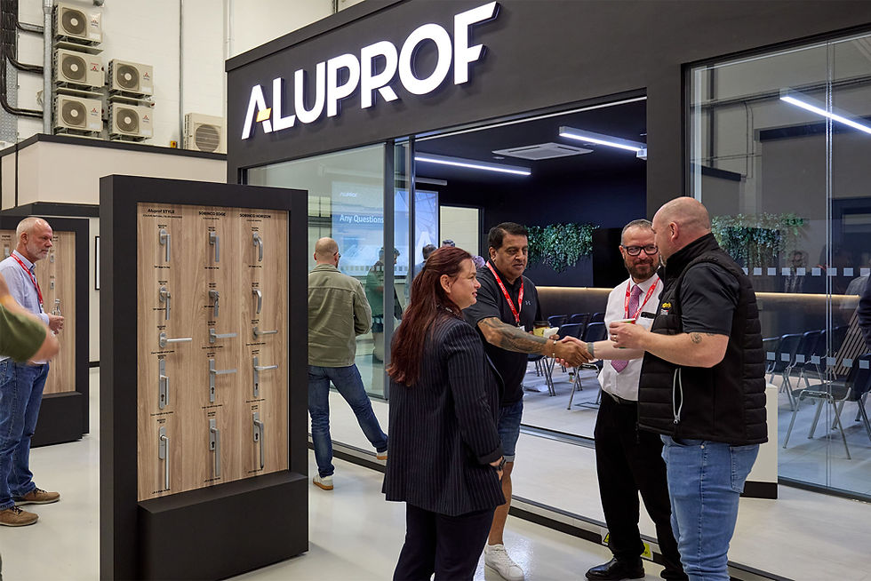 People in a showroom greet each other with a handshake. "ALUPROF" sign above. Display cases with hardware, indoor setting, casual attire.