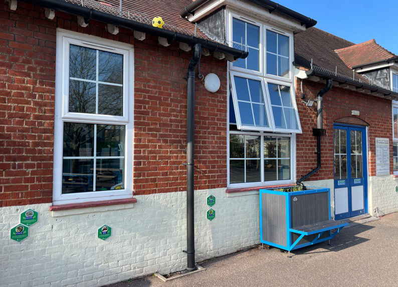 Red-brick building with white lower wall, blue-framed windows and door, a planter bench outside, and a yellow ball on the roof.
