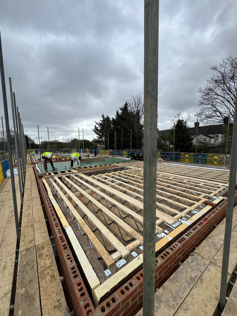 Construction workers install timber roof joists at a building site under a cloudy sky, with scaffolding and brick walls.