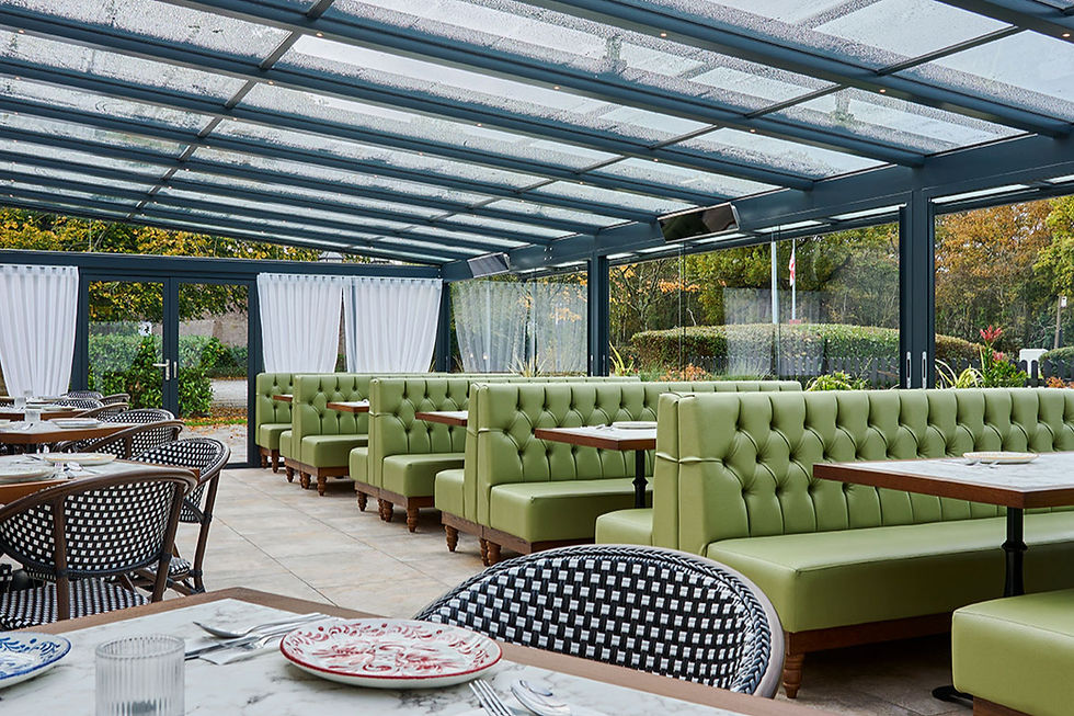 Empty modern restaurant with green banquettes, checkered chairs and set tables under a glass roof, with garden outside.