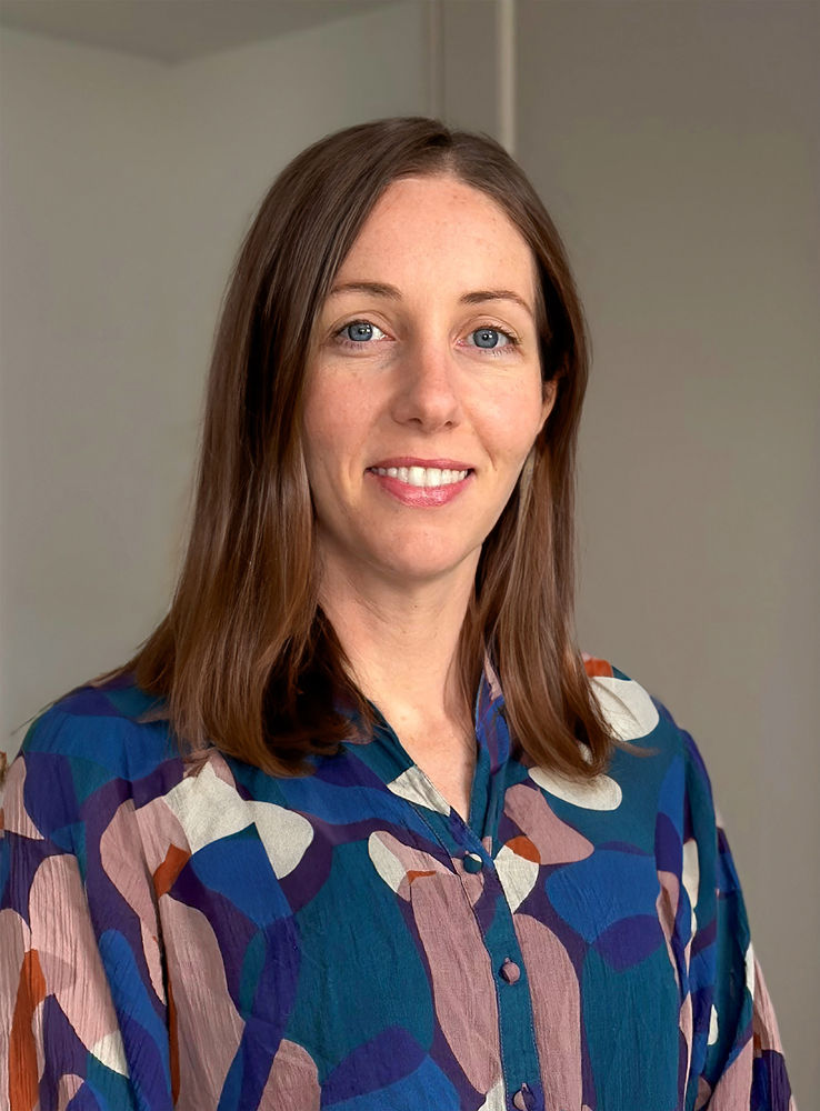 Woman smiling in a colorful, patterned blouse stands against a plain background. The mood is cheerful and welcoming.
