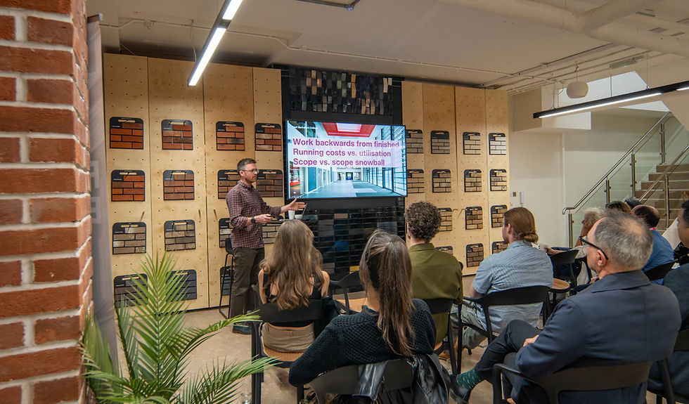 A man presents to seated audience in a modern room. Screen displays text on project management. Brick designs and plants adorn the space.