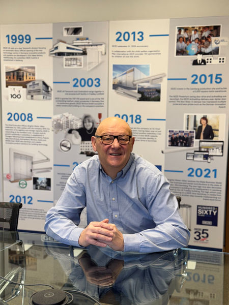 Smiling man sits at a glass table in front of a timeline display from 1999 to 2021, featuring corporate achievements and photos.