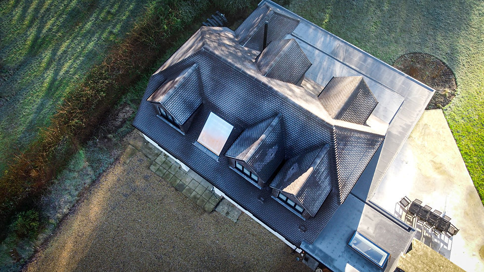 Aerial view of a dark-roofed house with dormers beside green fields, patio furniture on the terrace, and long morning shadows.