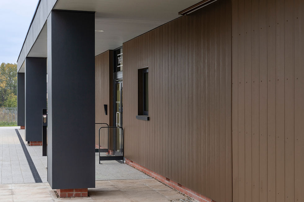 Modern building exterior with dark columns and brown panel walls. A glass door leads inside. Trees are visible in the distance.