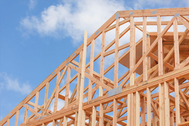 Wooden house frame under construction against a blue sky with clouds, showing exposed roof trusses and beams.
