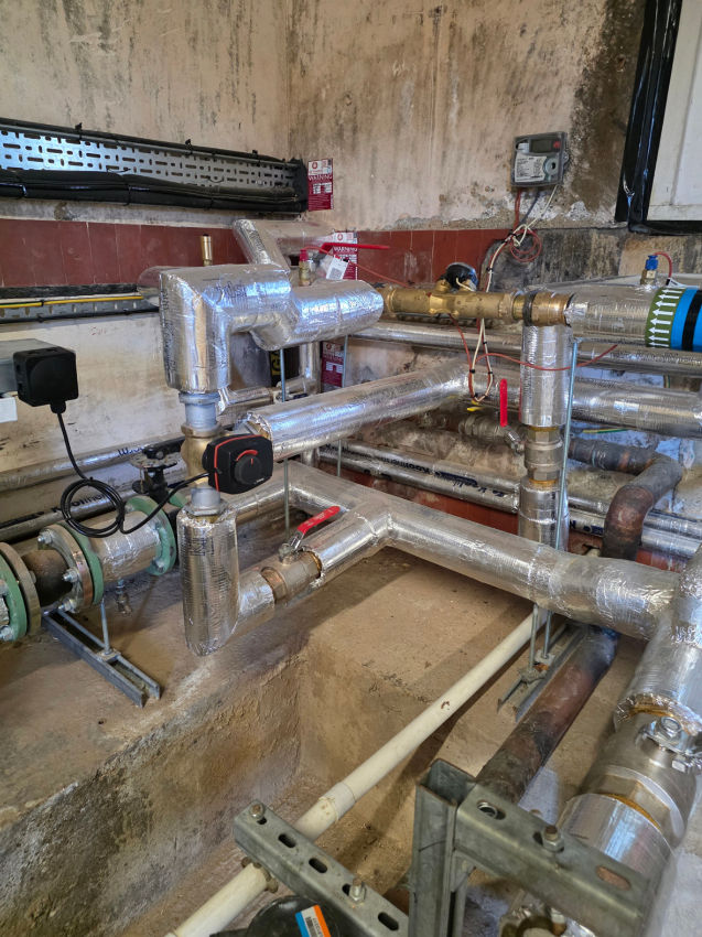 Insulated industrial pipes and valves in a worn mechanical room, with meters and wiring against stained walls.